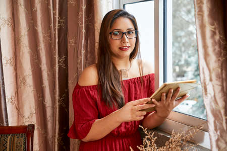 Pretty smiling young woman in digital table standing at window, she is working from home due to coronavirus pandemicの写真素材