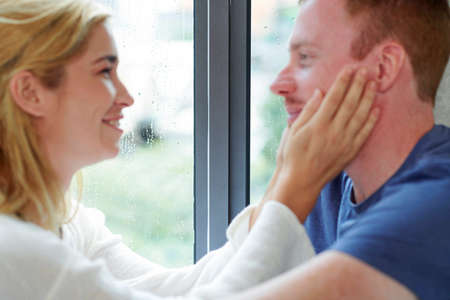 Young couple in love staying home on rainy day, they are touching faces and looking at each other, focus on window in backgroundの写真素材