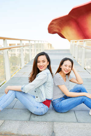 Beautiful young Vietnamese female friends sitting back to back on bridge and smiling at cameraの写真素材