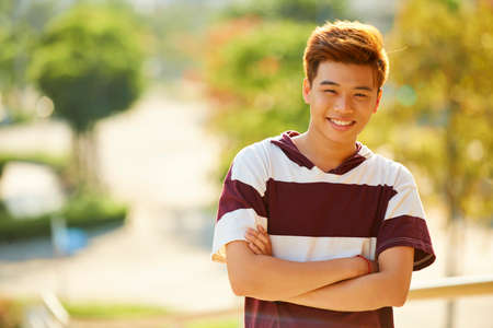Portrait of happy young man in striped t-shirt crossing arms and looking at camera when standing outdoors on summer dayの写真素材