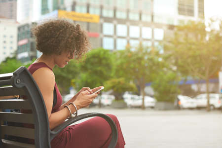 Pretty curly young woman sitting on bench outdoors and answering text messages of friends and colleaguesの写真素材
