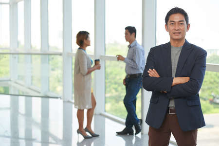 Portrait of confident smiling Vietnamese entrepreneur standing with arms crossed, his colleagues talking in backgroundの写真素材