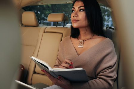Pensive young Asian businesswoman in necklace sitting in backseat and wrinting down her ideas in diaryの写真素材