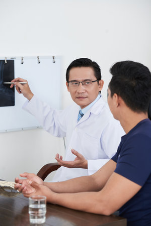 Vertical portrait of mature Asian doctor wearing white coat and eyeglasses sitting at desk showing X-ray shot to his patient explaining something to himの写真素材