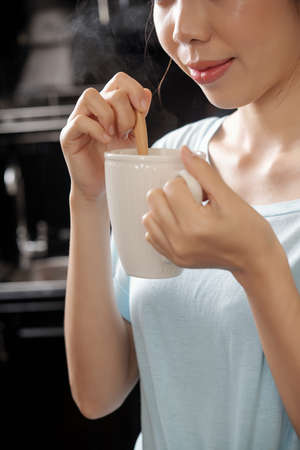 Close-up of content Asian girl stirrign sugar with spoon and smelling coffee scent in kitchenの写真素材