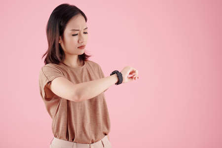 Frowning young woman reading notification on her smartwatch, isolated on pinkの写真素材