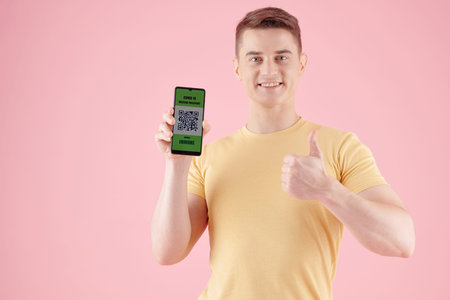 Portrait of excited smiling young man showing thumbs-up and smartphone with QR code after getting...