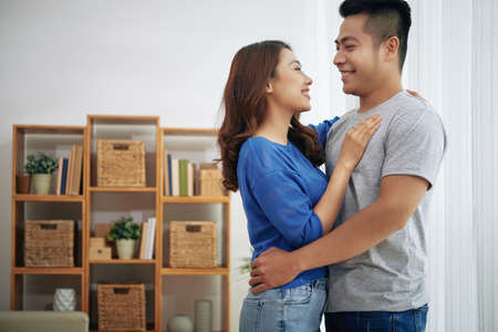 Smiling young Asian couple embracing while dancing in living room with shelves in backgroundの写真素材