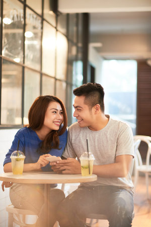 Smiling beautiful young Asian couple sitting at small round table with refreshing cocktails in cafe and scrolling through smartphoneの写真素材