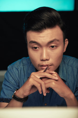 Focused young Asian man in wristwatch leaning head on hands and looking at monitor in dark room