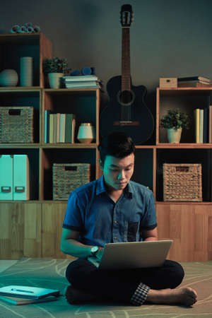 Busy young Asian marketer sitting with crossed legs on floor against shelves with guitar and working with laptop at nightの写真素材