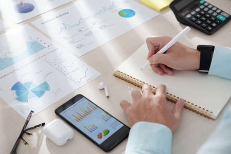 Hands of business person taking notes when working with charts on smartphone screenの写真素材