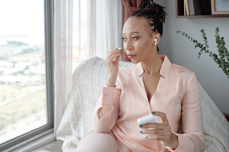 Gorgeous young woman wearing earbuds and looking through big window of her apartment