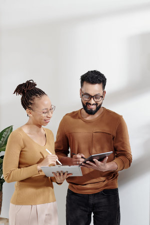 Happy young businessman and businesswoman with digital tablets exchanging ideas when standing in...