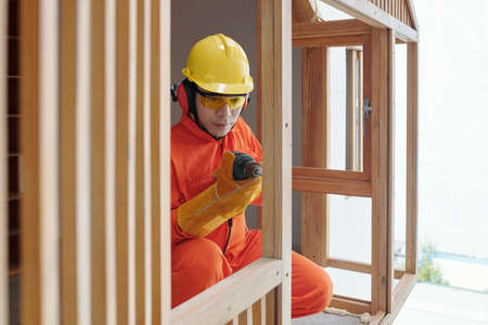 Serious builder in protective helmet, goggles and uniform building timber frameの写真素材