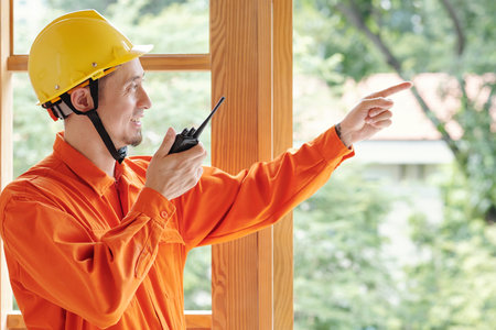 Smiling foreman talking on walkie-talkie when controlling work of his colleague at construction site