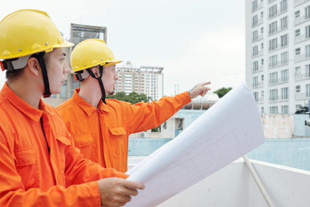 Construction workers standing at site with blueprint in hands and pointing at skyscraper near by