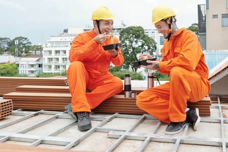 Team of smiling builders eating lunch and drinking coffee on rooftop after productive morning at construction siteの写真素材