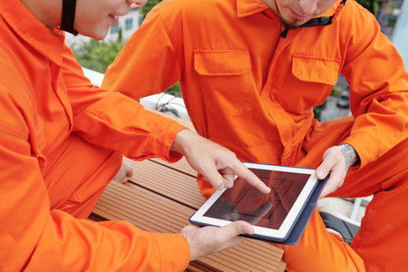 Closeup image of construction workers in bright orange overalls discussing blueprint on digital...