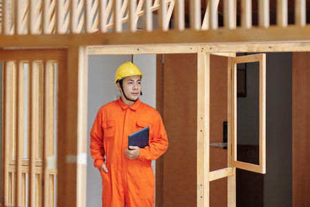 Foreman in orange overall and hardhat holding tablet computer and walking around house under...