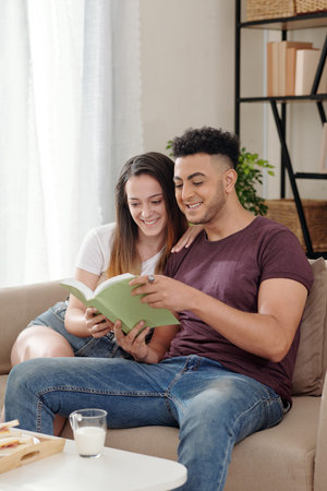 Cheerful young boyfriend and girlfriend sitting on couch with book in hands