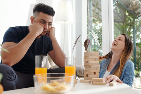 Pretty young woman laughing celebrating victory in board game when frustrated husband looking at...