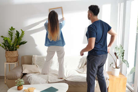 Young couple deciding where to hang picture frame on the wall in living roomの写真素材