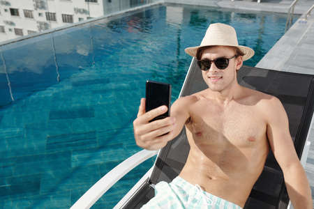Smiling young man taking selfie in hat and sunglasses when spending vacation by swimming poolの写真素材