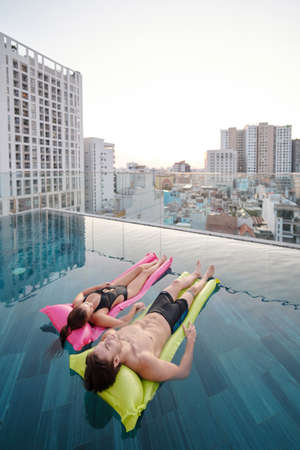 Boyfriend and girlfriend holding hands when relaxing in rooftop swimming pool with beautiful city viewの写真素材