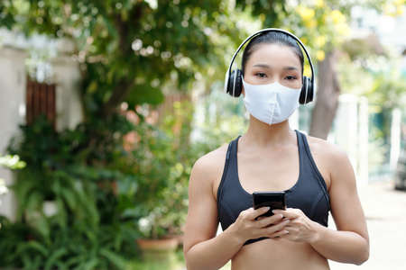 Portrait of attractive young Asian runner in sports clothe in protective mask and headphones holding smartphone and looking at cameraの写真素材