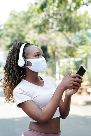 Young woman in medical mask walking outdoors and choosing what music track to listen to next
