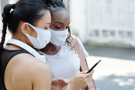Young woman in protective mask showing new sports mobile application to friend when they are...