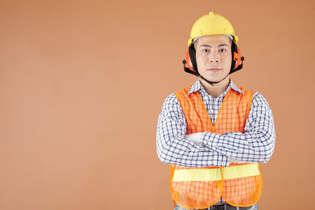 Portrait of construction worker in bright vest, hardhat and earmuffs crossing arms and looking at cameraの写真素材
