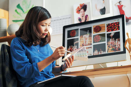 Concentrated young Asian woman in blue sweater using color swatch while choosing color palette for design of projectの写真素材