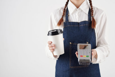 Coffeeshop barista holding disposable cup of coffee and payment terminalの写真素材