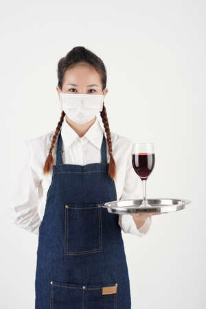 Portrait of positive young waitress in protective mask holding tray with glass of red wineの写真素材