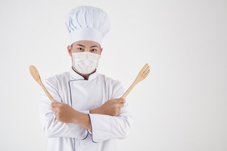 Portrait of cheerful chef in medical mask posing with wooden spoon and spatulaの写真素材