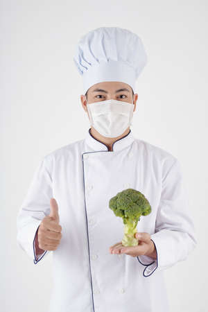Portrait of positive chef in protective mask holding fresh broccoli and showing thumbs-upの写真素材