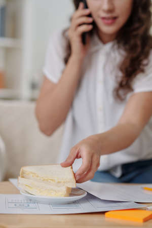 Young businesswoman eating sandwich when talking on phone with colleague and disccusing latest reportの写真素材