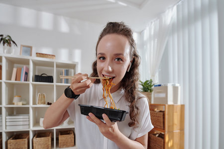 Cheerful young woman eating noodles for lunch in her home officeの写真素材