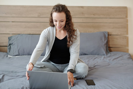 Positive young businesswoman sitting on bed with opened laptop and getting ready for online...