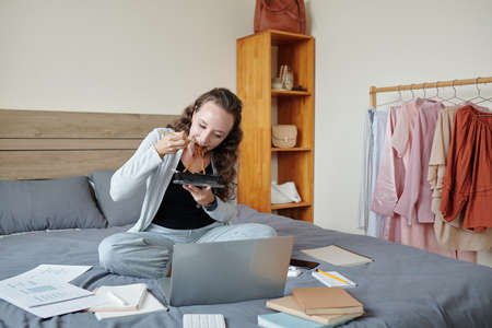 Young woman sitting on bed with documents around, eating lunch and reading documents and e-mails on laptop screenの写真素材