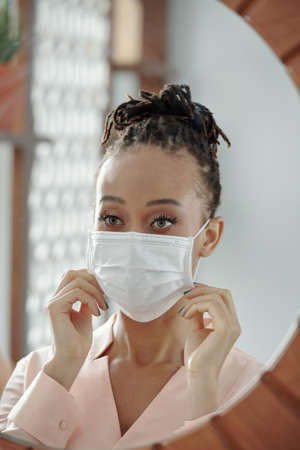 Gorgeous Black young woman with beautiful eyes looking at mirror and putting on protective maskの写真素材