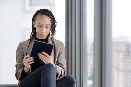 Pensive young woman sitting on window sill and drawing illustration on tablet computer with digital penの写真素材