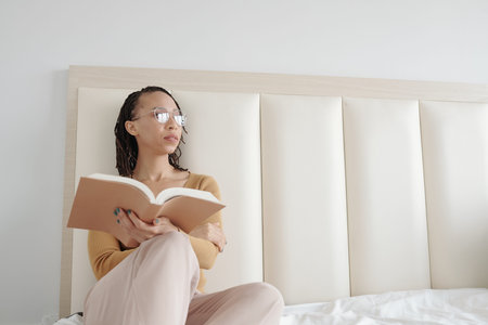 Pensive young woman in glasses sitting on bed and looking away after reading emotional scene in book