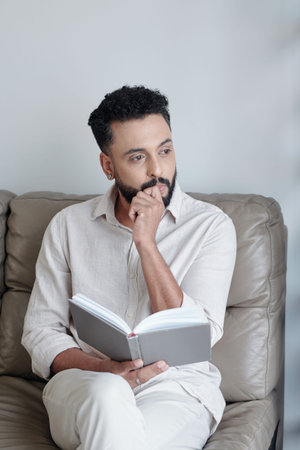 Contemplating man sitting on sofa at home with opened book and looking awayの写真素材