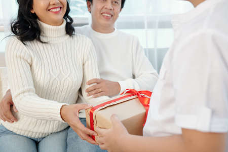 Child giving Christmas present to his happy parents sitting on sofaの写真素材