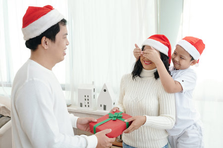 Smiling boy covering eyes of mother when dad giving her Christmas presentの写真素材