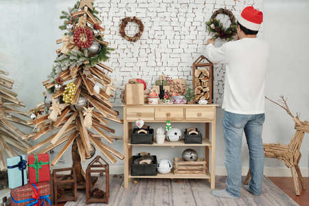 Man hanging handmade Christmas wreath on wall when decorating apartment for winter holidaysの写真素材