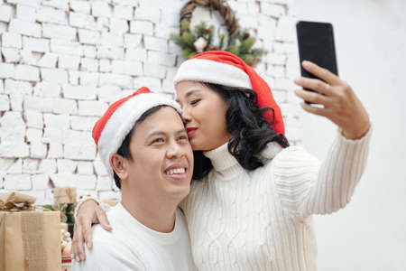 Pretty woman in sweater and santa hat kissing husband on forehead and taking selfie for social mediaの写真素材